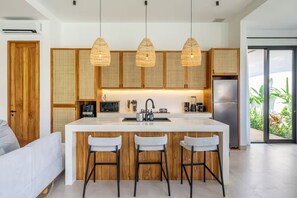 A modern kitchen with a central island, bar stools, hanging rattan light fixtures, and a view of greenery through a sliding glass door. - Julia Villa Bali (Gianyar)