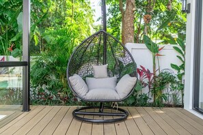 A hanging chair with a cushion in a garden area with plants and a wooden deck. - Julia Villa Bali (Gianyar)