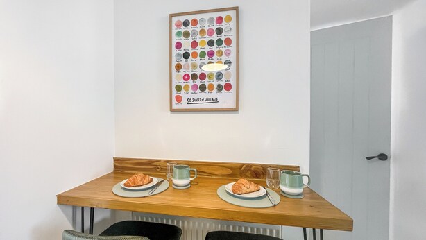 Een kleine eethoek met een houten tafel, twee stoelen, een lijst met een afbeelding van Schotse whisky en een deur op de achtergrond.