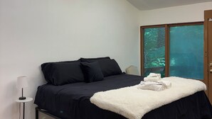 Un dormitorio con cama, lámpara de noche, lámpara de techo y ventana con vista a árboles.