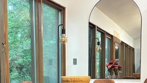 Un comedor con mesa de madera, sillas tapizadas en tono marrón, un espejo grande y vistas a zonas verdes a través de ventanas amplias.
