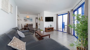 Ein modernes Wohnzimmer mit einer Couch, einem Couchtisch und einem Fernseher. Es gibt einen Essbereich mit Stühlen und einem Tisch, sowie ein großes Fenster mit Vorhängen.