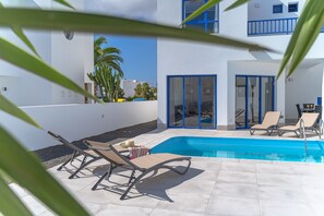 A poolside area with lounge chairs, a swimming pool, and a building with blue accents and glass doors. - Villa Yaiza 10 - VipVipVillas (Playa Blanca)
