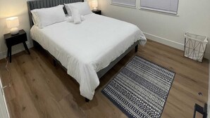 A bedroom with a bed, two bedside tables, a wall-mounted artwork, two windows with blinds, a patterned rug, and a waste basket.