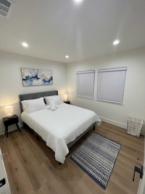 Un dormitorio con cama, dos mesitas de noche, un cuadro en la pared, dos ventanas con persianas, un tapete estampado y un cesto de basura.