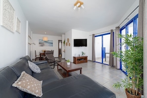 Ein modernes Wohnzimmer mit einer Couch, einem Couchtisch und einem Fernseher. Es gibt einen Essbereich mit Stühlen und einem Tisch, sowie ein großes Fenster mit Vorhängen.
