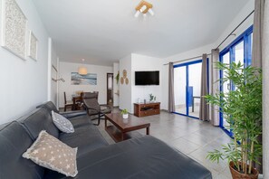A modern living room with a sofa, coffee table, and a television. There is a dining area with chairs and a table, and a large window with curtains. - Villas Yaiza 2 - VipVipVillas (Playa Blanca)