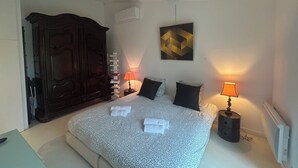 A bedroom with a bed, two bedside lamps, a wall-mounted air conditioner, a decorative wall piece, and a tall stack of books.
