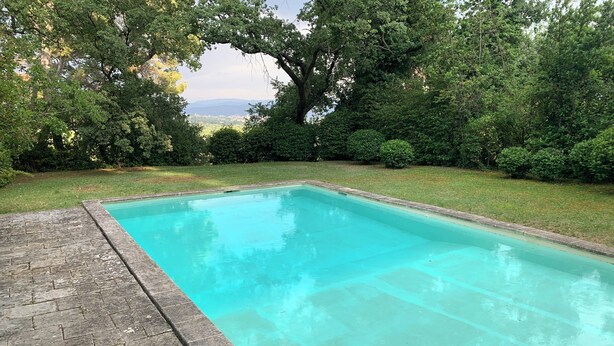 A rectangular swimming pool with a clear blue water surface, surrounded by a paved deck and lush greenery.