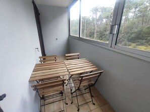 A balcony with a table and chairs, a window with a view of trees, and a tiled floor. - Proche commerces vue foret (Saint-Jean-de-Monts)