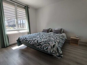 Une chambre à coucher avec un lit, une fenêtre avec des rideaux, un radiateur et une petite table de chevet en bois.