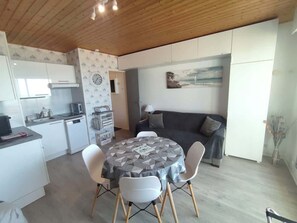 Una cocina moderna con armarios blancos, horno integrado y microondas. Hay un comedor con una mesa redonda y cuatro sillas. Un sofá gris está apoyado en la pared, y hay un cuadro encima.