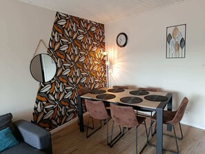 Un comedor con una mesa redonda, seis sillas, un muro con diseño de hojas, un reloj y una obra de arte enmarcada.