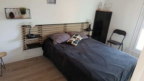 Una cama con cabecera de madera, una mesita de noche con lámpara, una silla y una estantería con objetos decorativos.