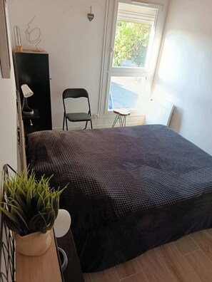 A bedroom with a bed, a chair, a small table, a potted plant, and a window with a view of greenery. - appartement cosy proche plage et commerces (Saint-Jean-de-Monts)