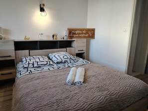 Un letto rifatto con cuscini decorati, una lampada da comodino, una piccola mensola con oggetti decorativi e un cartello motivazionale al muro.