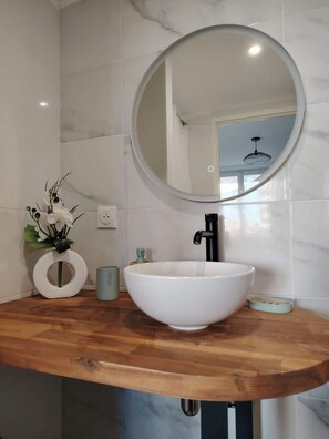 A bathroom with a wooden vanity, a white sink, a round mirror, and a vase with white flowers. - Belle chambre accès plage (Saint-Jean-de-Monts)