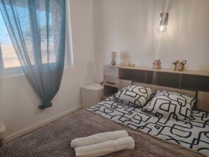 Una camera da letto con un letto, un comodino, una lampada, uno scaffale con oggetti decorativi e una finestra con tende leggere.