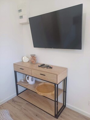 A room with a TV mounted on the wall, a wooden console table with drawers, a shelf, and a basket. - Belle chambre accès plage (Saint-Jean-de-Monts)