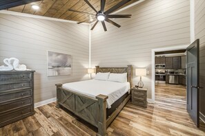 Une chambre avec un lit en bois, une commode, une table de chevet avec une lampe, un ventilateur de plafond et une vue sur la cuisine.