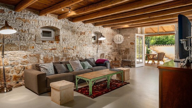 A rustic interior with a stone wall, wooden beams, a sofa, a coffee table, and a view through a glass door. - Villa Eva Rustica (100591-K1) - Zbandaj (Zbandaj)
