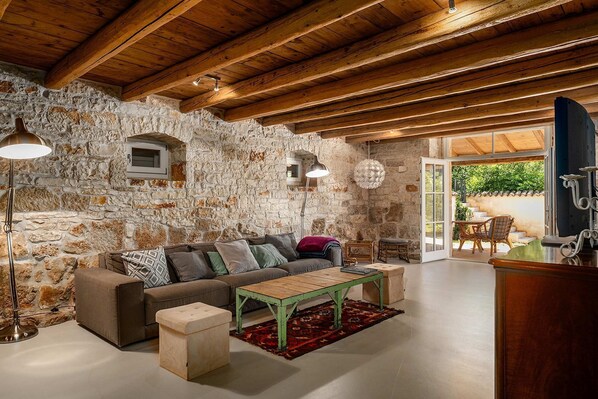 Ein rustikaler Innenraum mit Steinwand, Holzbalken, einer Couch, einem Couchtisch und Blick durch eine Glastür.