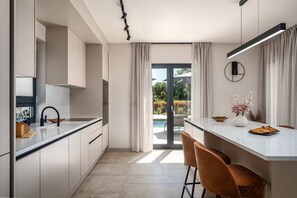 Eine moderne Küche mit einer großen Kochinsel, einem Essbereich mit Stühlen und Blick ins Freie. An der Wand hängt eine Uhr.