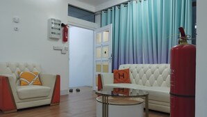 A living room with a white sofa, a round table, a red fire extinguisher, and a blue curtain. - TRANSIT HOTEL  LUXUARY (Male)