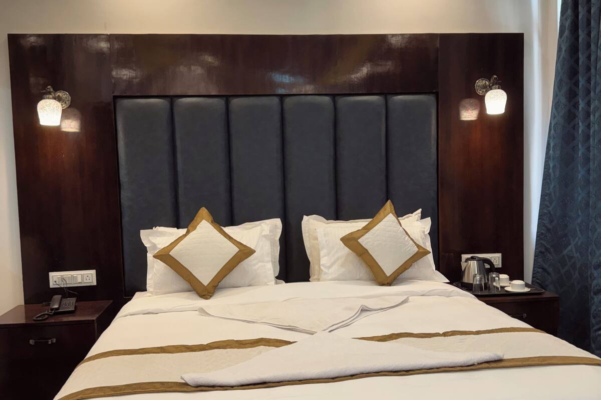 A hotel room with a large bed, dark headboard, bedside lamps, and a nightstand with a phone and a kettle.