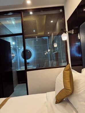 Quarto de hotel com cama, janela com vista para um elemento arquitetônico circular, área de chuveiro e luminária fixada na parede.