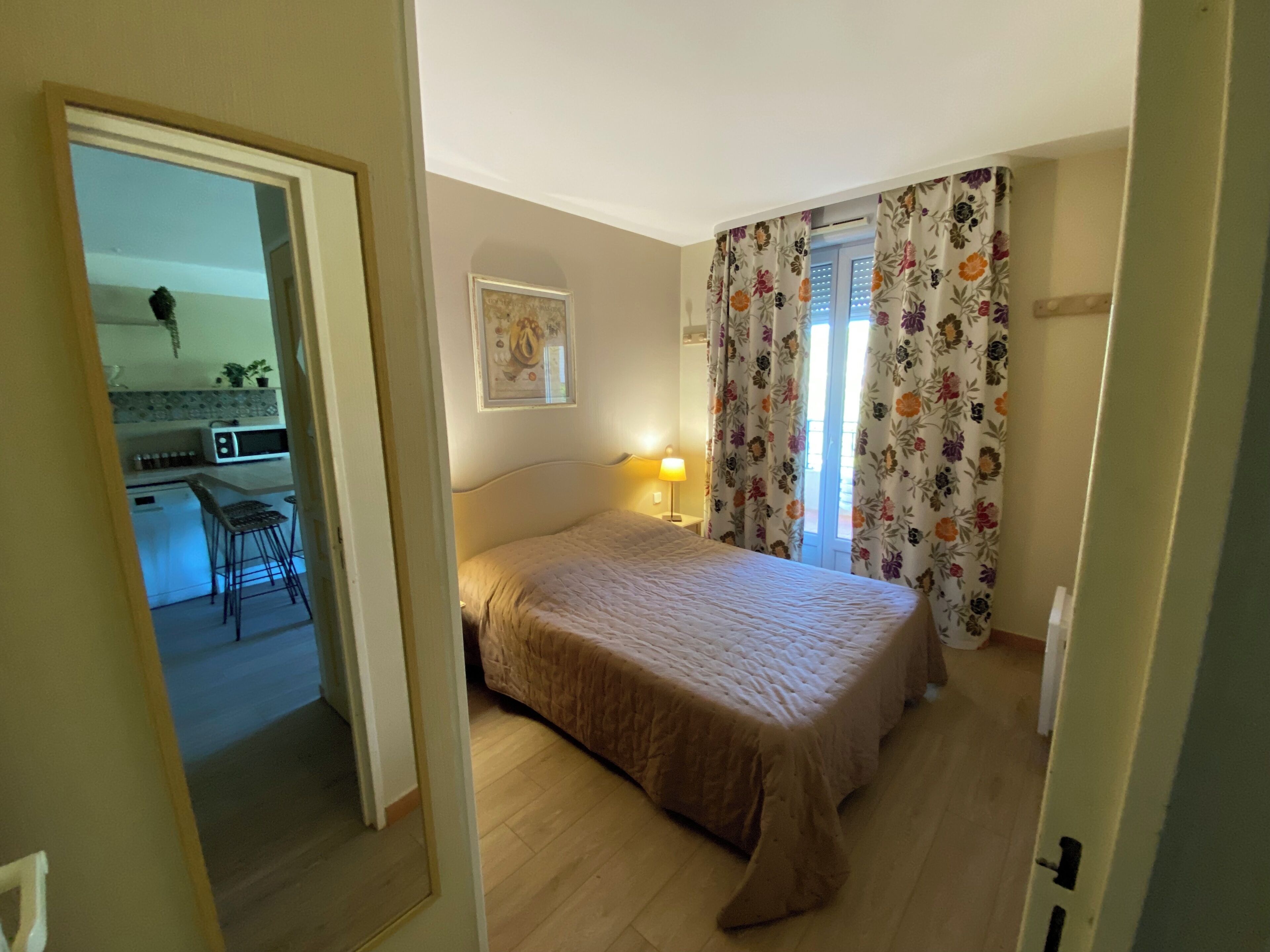 A bedroom with a bed, a nightstand, a lamp, a window with curtains, and a view of a kitchen area.