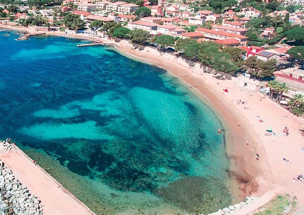 An aerial view of a coastal area with clear turquoise waters, a sandy beach, and a densely built residential area with red-roofed houses.