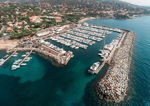 An aerial view of a marina with numerous boats docked, a rocky breakwater, and a residential area in the background.