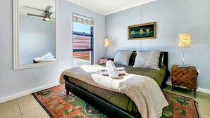 Ein Schlafzimmer mit einem Bett, einem Deckenventilator, einem Fenster mit Jalousien, einem Bild, einem Nachttisch mit Lampe und einem gemusterten Teppich.