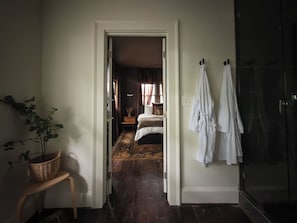 Ein Flur, der zu einem Schlafzimmer führt, mit einem Bett, einer Topfpflanze, einem Korb aus Weidengeflecht und einer Duschkabine aus Glas.