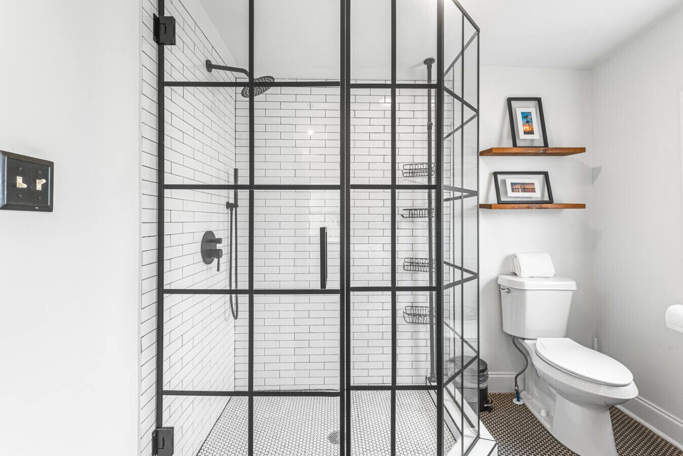 A modern bathroom with a glass shower enclosure, white tiled walls, a toilet, and a shelf with framed pictures.. 5 min to Mercedez Benz | Front Porch | Workspace
