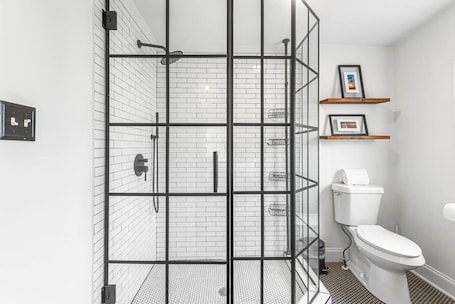 A modern bathroom with a glass shower enclosure, white tiled walls, a toilet, and a shelf with framed pictures.. 5 min to Mercedez Benz | Front Porch | Workspace