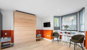 A modern room with a wooden sliding door, a TV mounted on the wall, a dining table with chairs, and a window with blinds. - Boutique Studio - Near Kensington & Notting Hill (London)