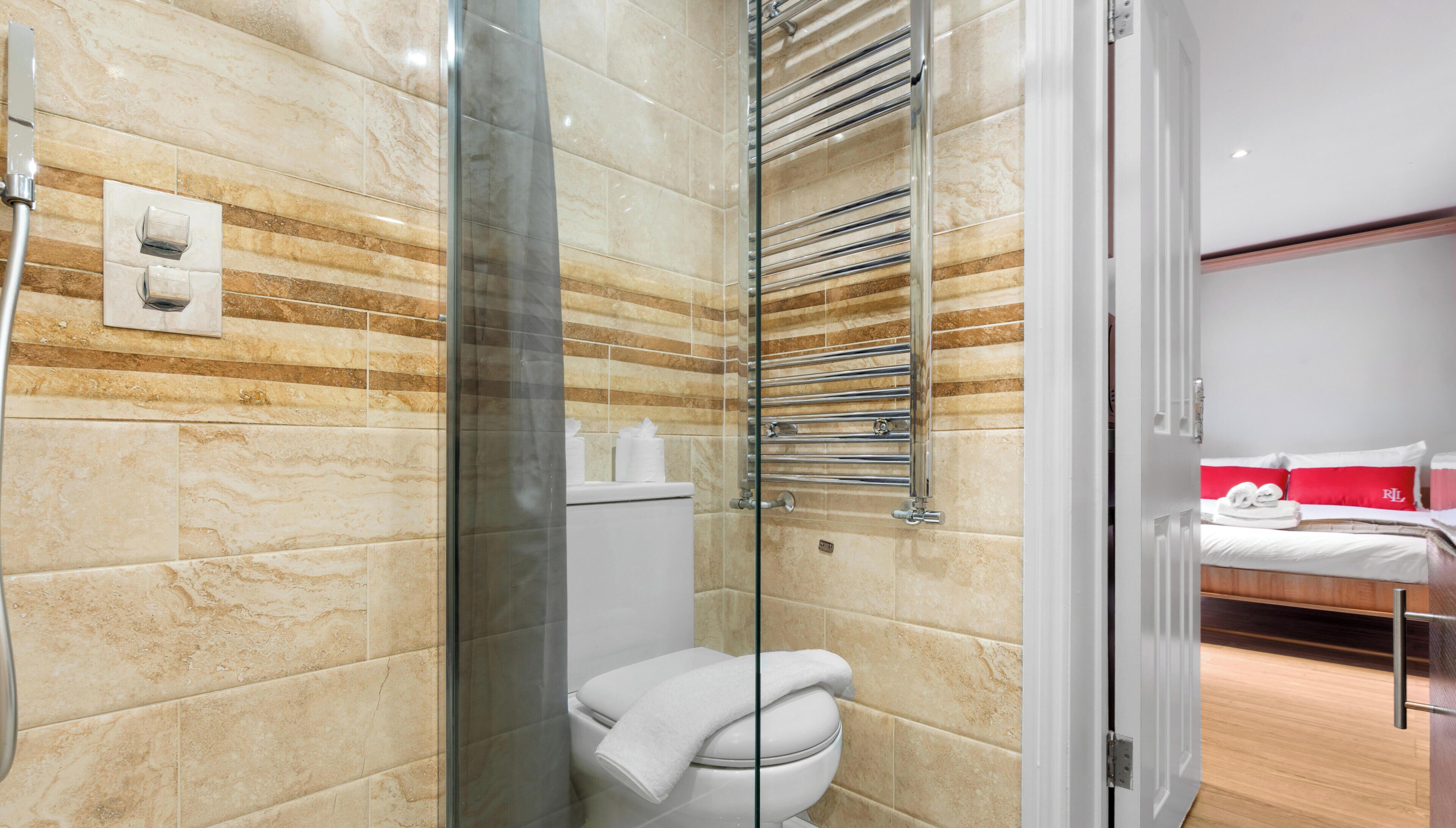 A modern bathroom with a glass shower enclosure, a toilet, and a glass door leading to a bedroom with a bed and a red pillow.