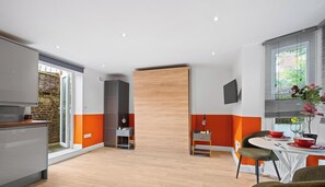 A modern kitchen with a sliding wooden door, a dining area with a table and chairs, and a large window with blinds. - Boutique Studio - Near Kensington & Notting Hill (London)