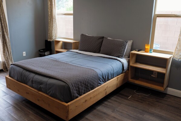 Habitación individual con cama de madera, colcha acolchada gris, mesita de noche con lámpara y ventana con cortinas.