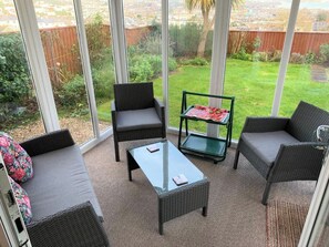Eine Terrassenfläche mit Korbmöbeln, einem Glastisch und Blick auf eine grüne Rasenfläche und einen Holzzaun.