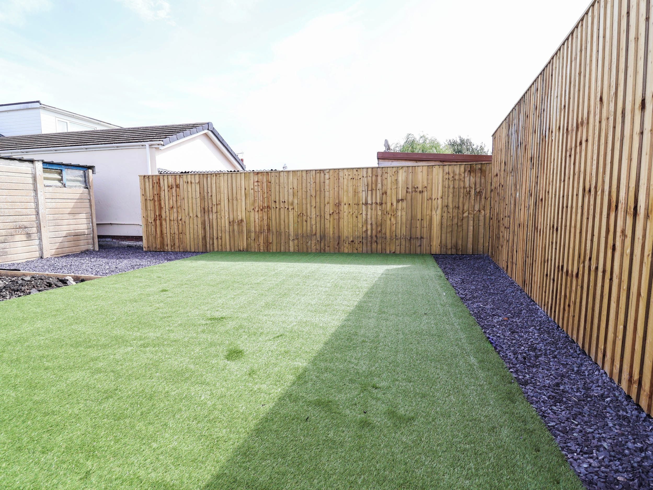A backyard with artificial grass, a wooden fence, and a gravel path.
