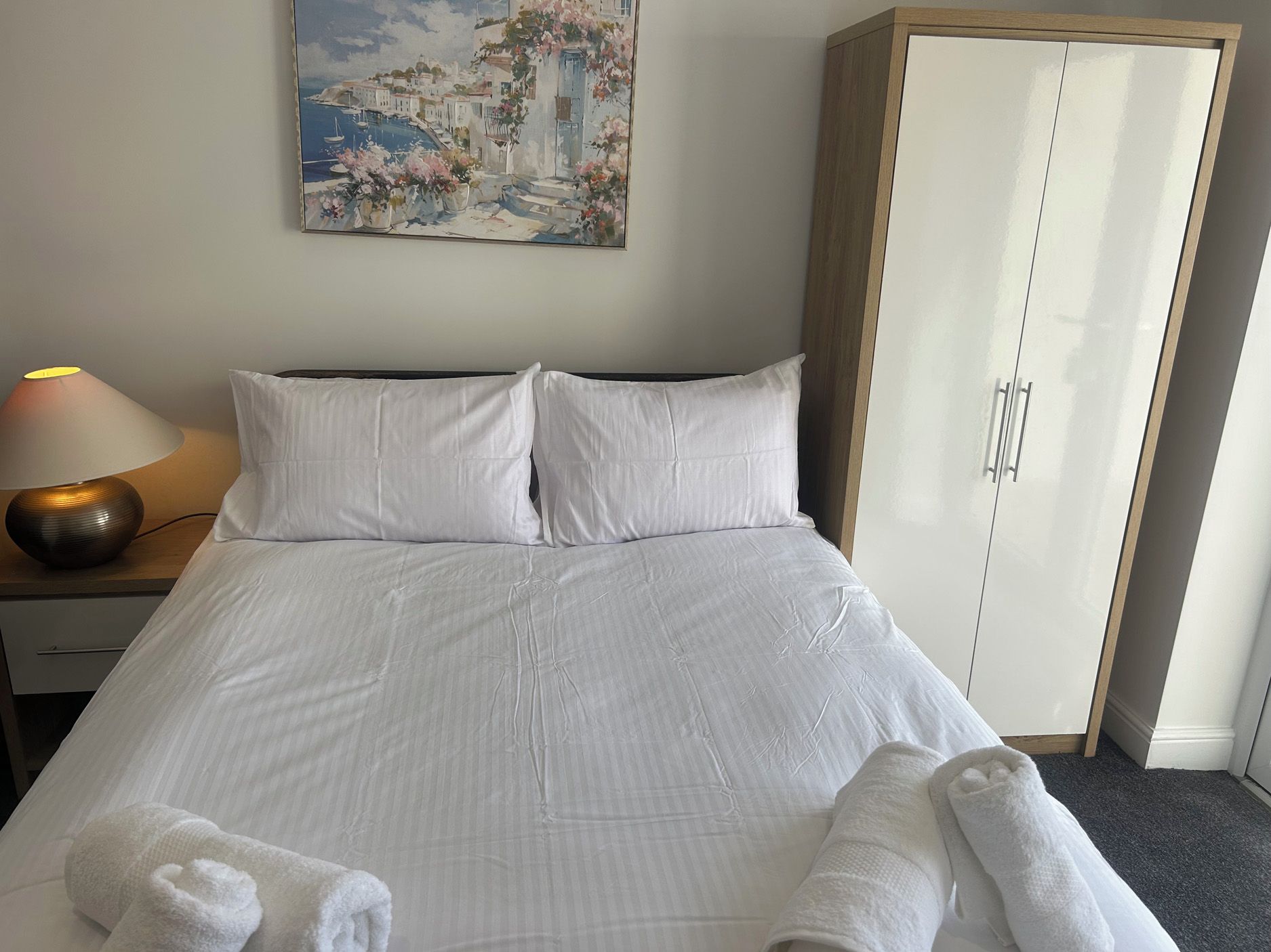 A hotel room with a bed, a nightstand with a lamp, a wardrobe, and a painting on the wall.