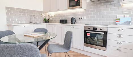 A modern kitchen with white cabinets, a glass dining table, grey chairs, a microwave, a coffee maker, and an oven.