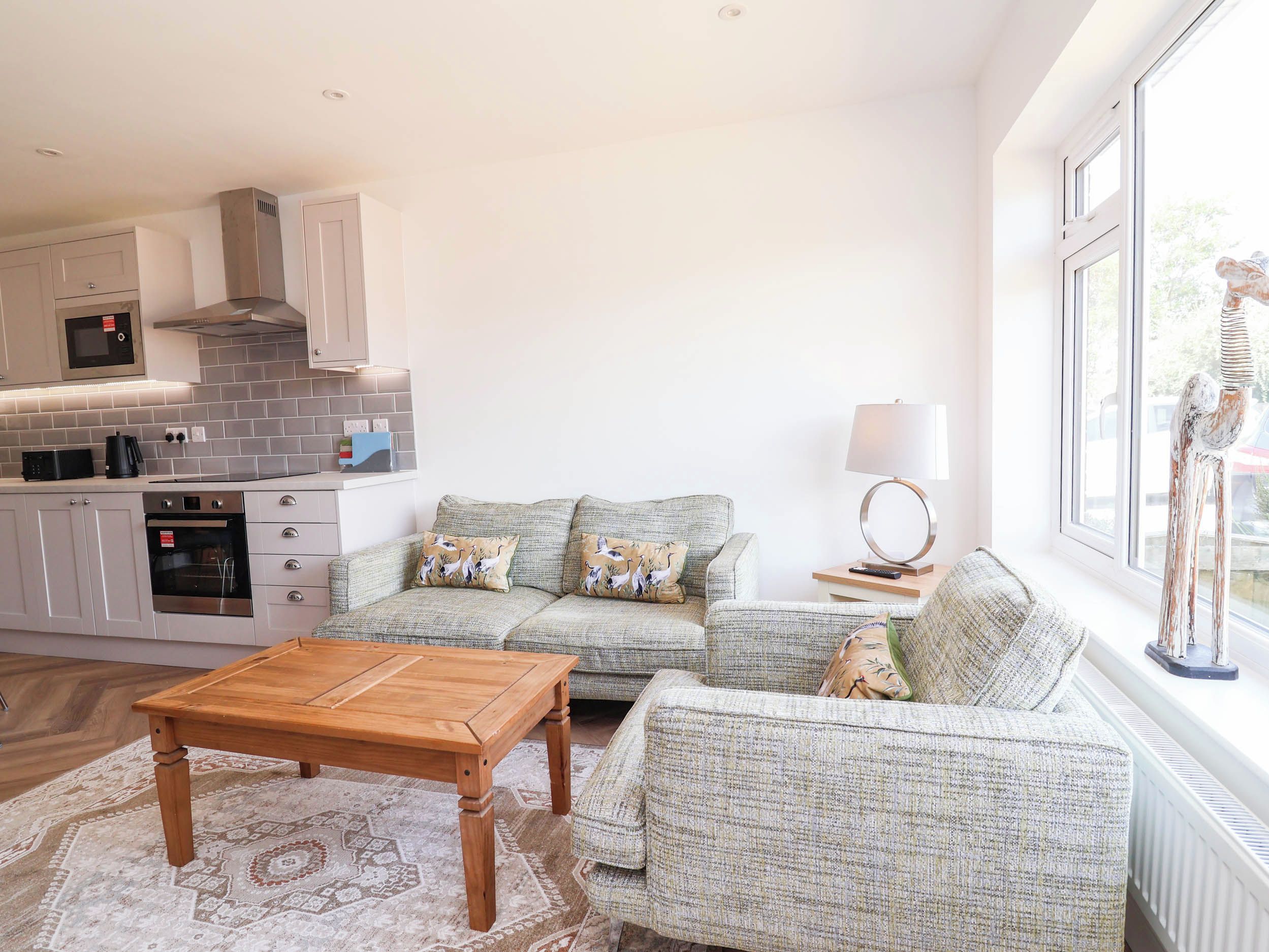 A modern kitchen with a built-in oven and microwave, a grey sofa with patterned cushions, a wooden coffee table, and a view of the outdoors through a large window.