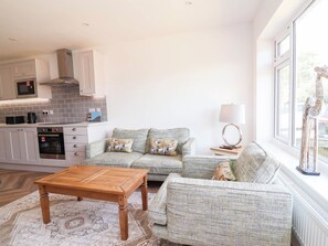 A modern kitchen with a built-in oven and microwave, a grey sofa with patterned cushions, a wooden coffee table, and a view of the outdoors through a large window. - 6 Bastion Close (Prestatyn)