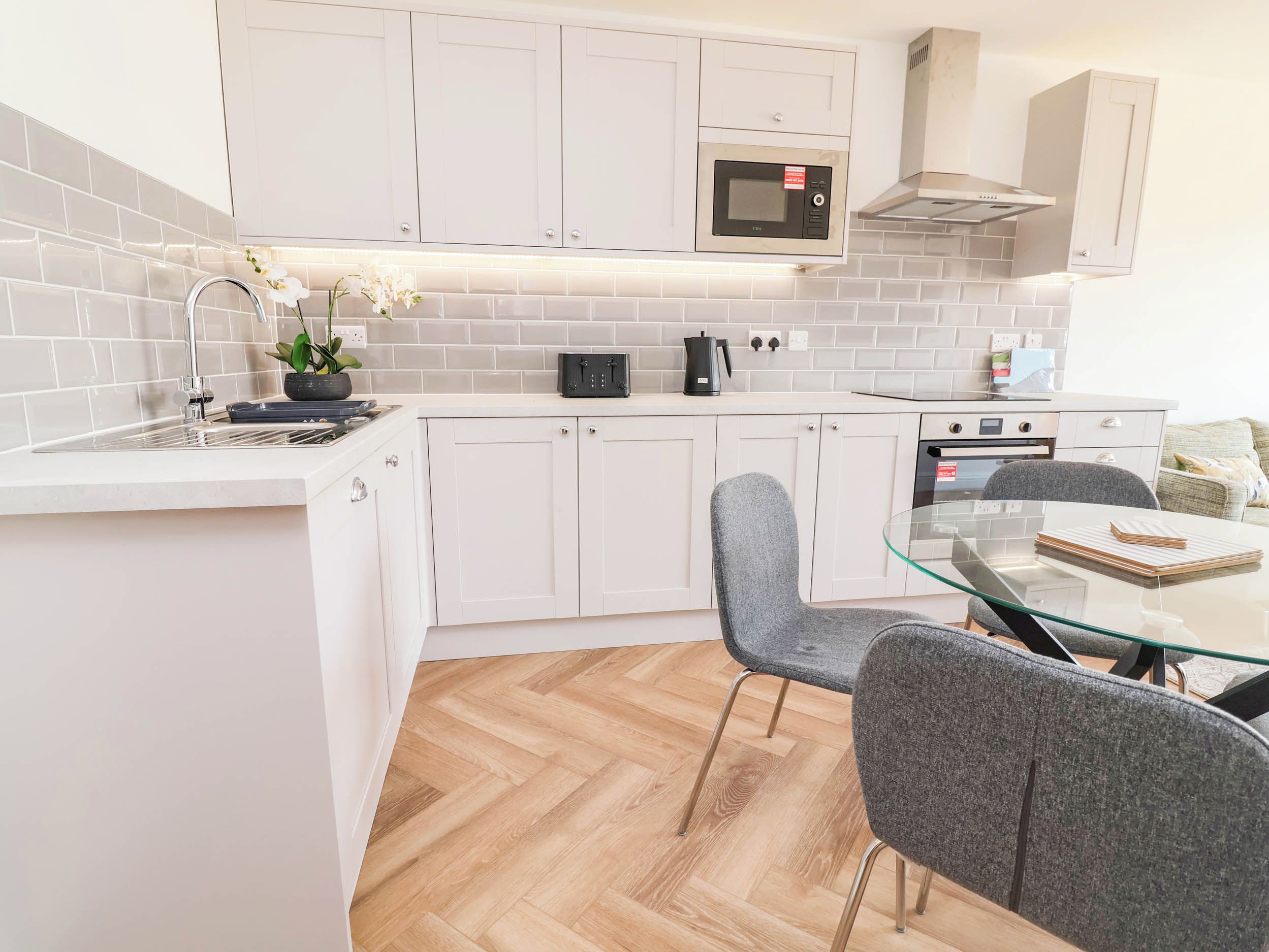 A modern kitchen with white cabinets, a microwave, a kettle, and a glass dining table with chairs.