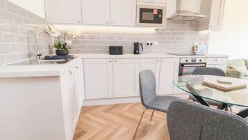 A modern kitchen with white cabinets, a microwave, a kettle, and a glass dining table with chairs.