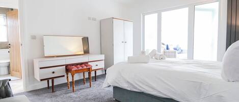 A modern bedroom with a large bed, a vanity with a mirror, a wooden bench, and a view of the outdoors through a large window.